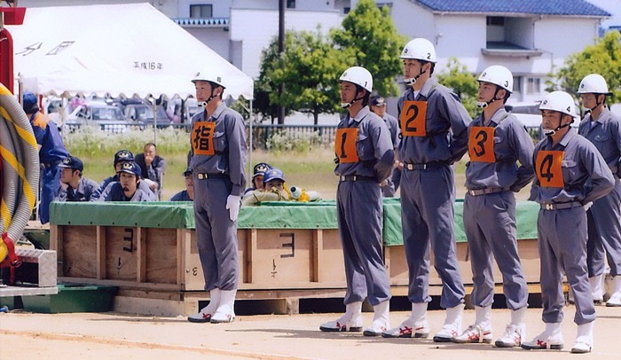 運動場のような場所で、数人の帽子をかぶった男性が見ている中で、ヘルメットをかぶりゼッケンを付けた男性5人ほどの男性が後ろに手を組んで横一列に並んでいる様子の写真