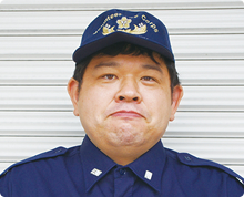 堂々とした顔つきの中嶋 靖博さんが高岡市消防団のキャップをかぶりシャッター扉の前で正面を向いている様子の写真