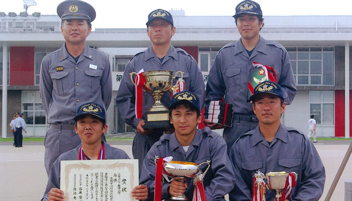 野外で、6人の消防団員が前列3人・後列3人横並びに並んでおり、前列の3人は賞状やトロフィーを手に持っており、後列の3人のうち2人はトロフィーを手に持っている様子の写真
