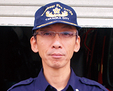 半分ほど開いたシャッター扉の前でメガネをかけた脇本 康史さんが高岡市消防団のキャップをかぶり真っ直ぐ正面を向いている様子の写真