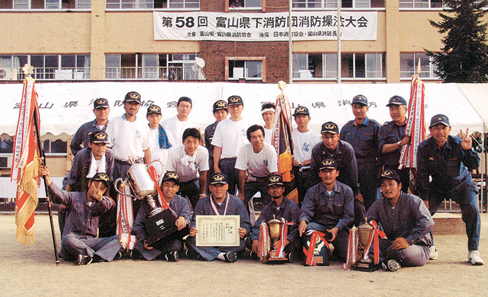 後ろに第58回富山県下消防団消防操法大会と書かれた垂れ幕のようなものがあり、その手前にテントのようなものがあり、その手前に三列に並んだ消防帽子のような帽子を被った複数の人達がトロフィーや賞状、旗を手に持っている様子の写真