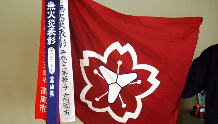 大きな旗に、5枚の花びらをかたどった図の中央に太いY字型の図が重なった形の消防団のマークが書かれており、無火災表彰・平成21年授与・高岡市などと書かれたペナントリボンが旗の左側に3本ついている様子の写真