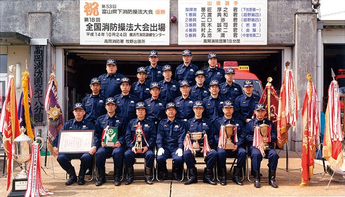 後ろに祝第18回全国消防操法大会出場などと書かれた看板のようなものの右隣に出場選手の6名の名前が書かれた看板があり、その手前に4列に並んだ消防帽子のようなものを身に纏った複数の男性らが賞状やトロフィーを手に持っておりその周りを囲むように、両端に優勝旗やトロフィーが幾つか並んでいる様子の写真
