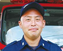 消防車の前で、大久保 慎一さんがキャップをかぶり消防団の制服を身に着け少し微笑んでいる様子の写真
