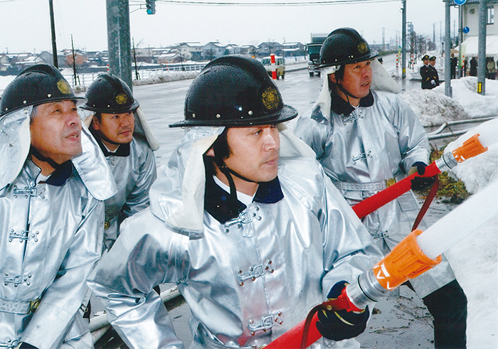 歩道などに雪が積もっている中、消防服を着た4人の男性が、2本のホースで放水作業を行っている様子の写真