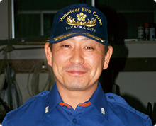高岡市消防団のキャップと制服を身に着けた中尾 徳志さんが正面を向いて微笑んでいる様子の写真