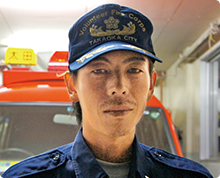 キャップをかぶった眉毛の凛々しい加藤 正和さんの後ろに消防署の車がある様子の写真