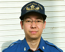 シャッター扉の前で、高岡市消防団のキャップと制服を身に着けメガネをかけた吉澤 満さんがやや微笑んで正面を向いている様子の写真