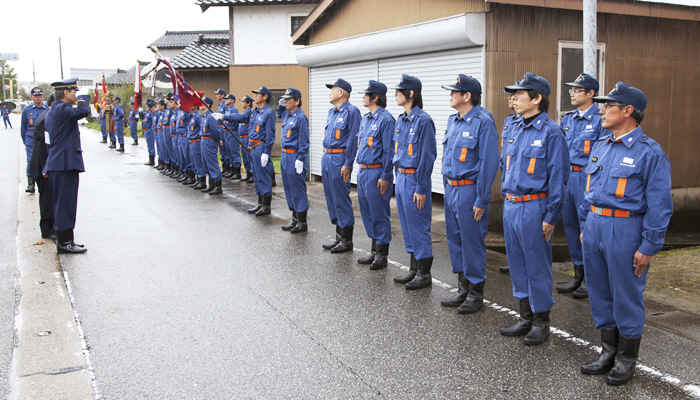 民家沿いの道路で敬礼する2名の男性と、それに向かい一列に整列している20名ほどの消防団員の様子の写真