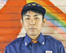虹のような絵が描かれたシャッター扉の前で、消防団のキャップと制服を身に着けた中村 卓哉さんが正面を向いている様子の写真