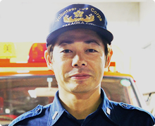 消防車の前で高岡市消防団のキャップをかぶり少し微笑んだ様子の林 靖浩さんが正面を向いている写真