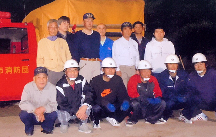 消防団の車の前で、3列に並ぶ13人の男性がおり、前列の男性はヘルメットを被ってしゃがんでおり、その奥の男性は立って並んでいる様子の写真