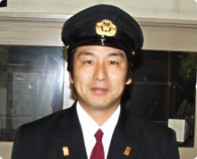 バッジのついた消防の制服を着てネクタイを締めている和泉 孝さんの後ろに窓がある様子の写真