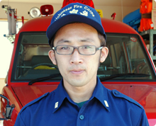 消防車の前で帽子をかぶって襟にバッジのついた服を着ている眼鏡をかけた大川 豊博さんの写真