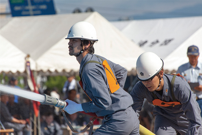 日よけテントが並ぶ野外で、ヘルメットをかぶりゼッケンを付けた男性が、2人共同で消防用のホースを使い放水作業を行っている様子の写真