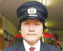 車庫のような場所で、凛々しい顔つきの消防団の高橋 克明さんが制帽をかぶり警備服を着て正面を向いている様子の写真