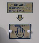 白い壁に貼られた、金色の長方形型の静電気除去シートと、シートに触れるよう促す注意書きの写真