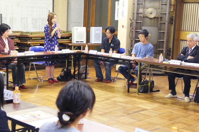 1人の女性がマイクを持ち立って話しているのを、参加者が聞いている様子を写した写真