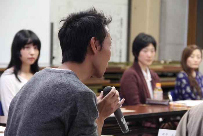 1人の男性がマイクを手に話しているところを参加者が聞いている様子を写した写真
