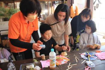 テーブルの前に座って菅笠づくりをしている少年の後ろに二人の女性が立ち、作業を手伝っている写真