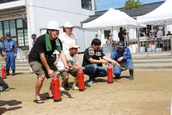 消防署の職員に指導を受けながら赤い消火器をそれぞれ持った男性3人が消火器を構えて消火訓練をしている写真