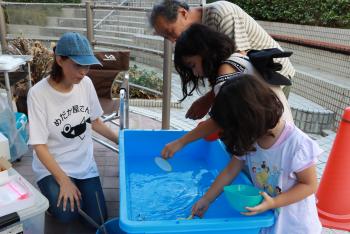 青い水槽の上にかがみこんで、めだかをすくおうとしている家族連れの写真