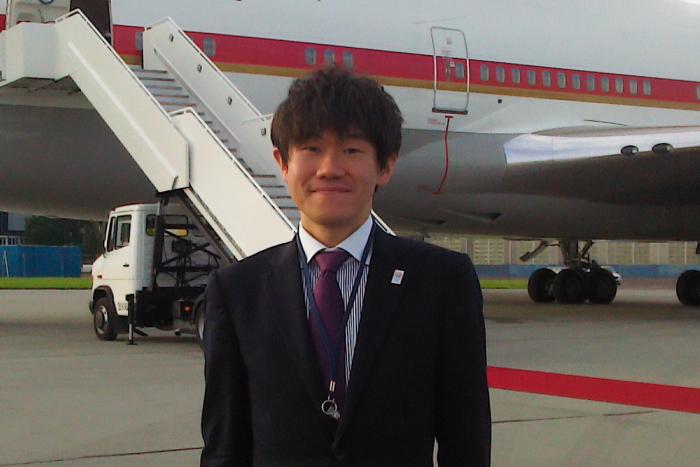 飛行機の乗降口の前で、スーツを着用して立つ室谷政克さんの写真
