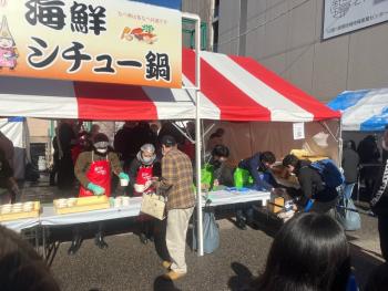 「海鮮シチュー鍋」と書かれた看板が立てられたテントで商品の用意をしている店員と出来上がりを待つ男性客の写真