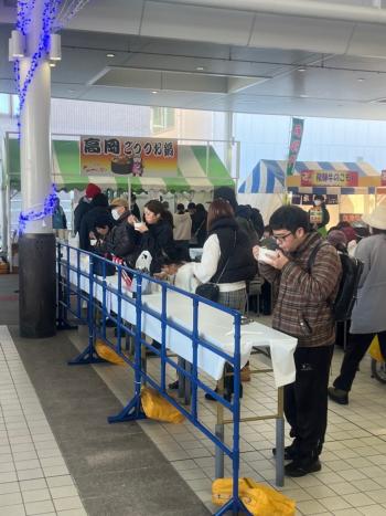 「高岡ごっつ鍋」と書かれた看板があるテント前の飲食スペースで美味しそうに食べている人たちの写真