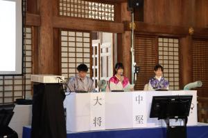 万葉衣装を着た男子生徒1人と女子生徒2人が、勝興寺本堂で、白いクロスをかけられたテーブルに座り、短歌バトルを行っている様子の写真
