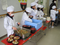 白い割烹着を着て配膳をしている給食当番の子どもたちの写真
