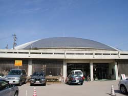 青空の下灰色のコンクリートの上に建てられているドーム状の建物の前に車が駐車している写真