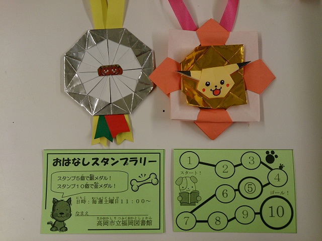 折り紙で作られた金と銀のメダルと、おはなしスタンプラリーのスタンプカードの写真