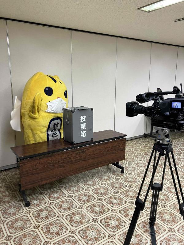 マスクを着用して投票箱と共に撮影機材の前に立つ、めいすいくんの写真