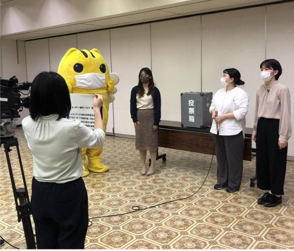投票箱の前で撮影に参加しているマスクをしためいすいくん、女性3名とカンペを出している女性の写真