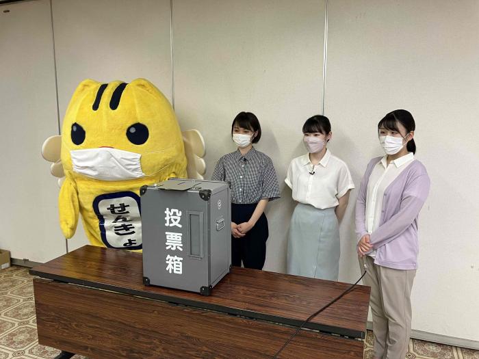 投票箱を前にして撮影に参加しているマスクをしためいすいくんと女性3名の写真