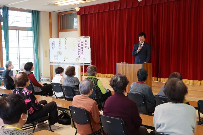 赤い幕が閉まったステージ前の演説台でしゃべるスーツ姿の男性と話を聞く年配の人達の写真