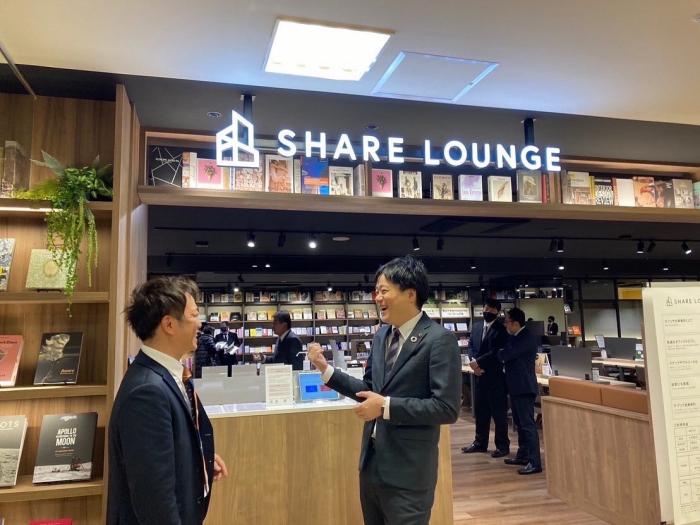 「SHARE LOUNGE」と書かれたネオン看板のついた本屋さんの前で笑顔で会話する二人のスーツ姿の男性の写真