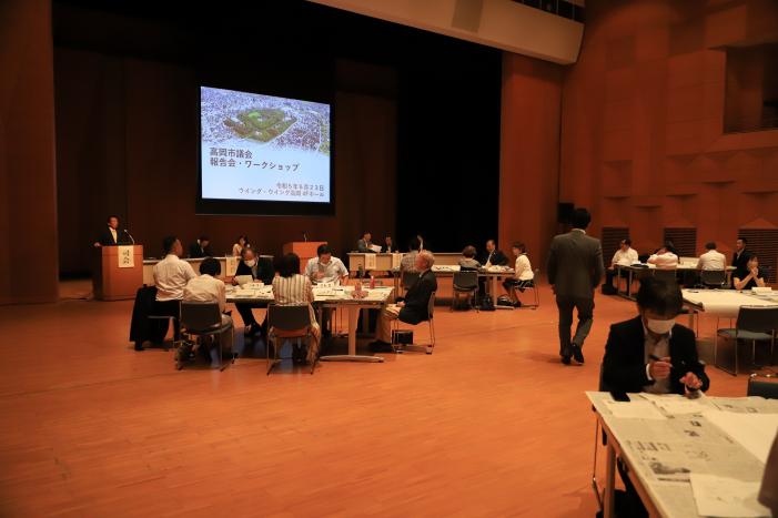 プロジェクターを前にして各グループに分かれ机に着席する議会報告会参加者たちの写真
