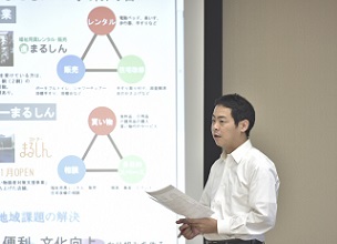 大きなスクリーン横で手に資料を持ちながらプレゼンテーションをしている塚田 高史さんの写真