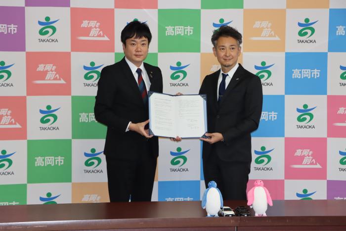 高岡市のロゴが入った衝立の前で、ネッツトヨタ富山株式会社と高岡市の代表者が、報道関係者に向かって協定の証書を広げて見せている写真