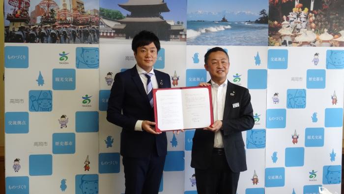 高岡市の様々な風景の写真が貼られた衝立の前で、二人の男性が、一冊の協定証書を開いて、報道陣に見せている写真
