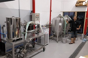 ビール工房内のビール製造装置の横でポーズを取る大島 紀明さんの写真