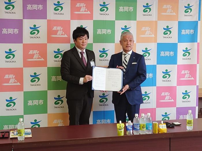 高岡市のロゴが入った衝立の前で、高岡市と大塚製薬の代表者が、協定の証書を報道陣に向かって広げて見せている写真