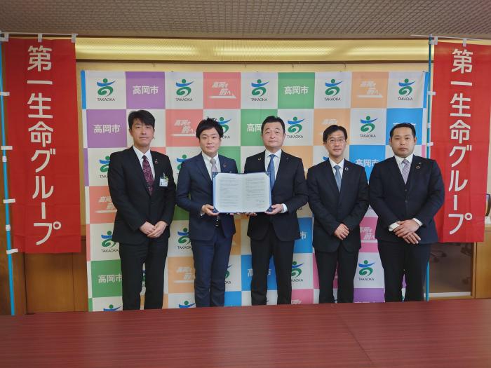高岡市のロゴが入った衝立と、第一生命グループと書かれた赤いのぼりの前で、五人のスーツ姿の男性が立ち、そのうちの二人が、一冊の協定の証書を一緒に開いて、報道陣に見せている写真