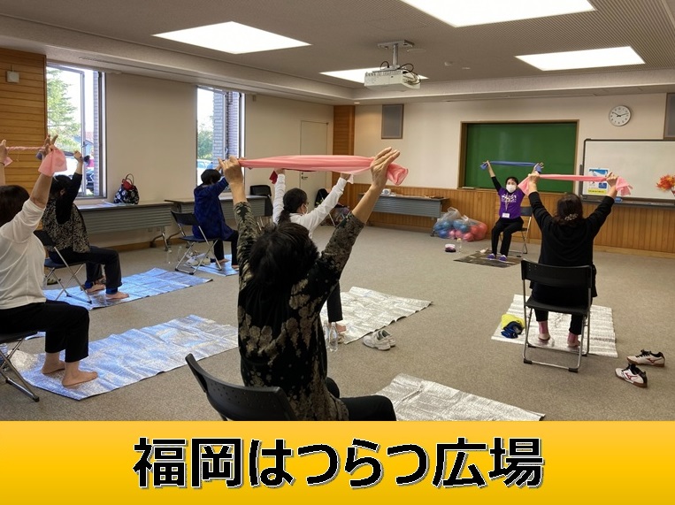 福岡はつらつ広場と書かれたタイトル書きと、複数の人々が手前に座ってタオルを掲げている女性を見ながら、椅子に座りタオルを上に挙げている様子の写真