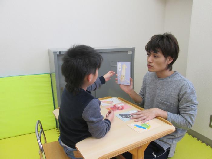 机に座った男の子にイラストカードを見せる男性の写真