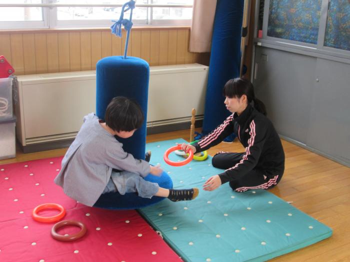 赤と水色のマットの上で、柱の付いたクッションに捕まっている子供に女性が投げ輪を渡している写真