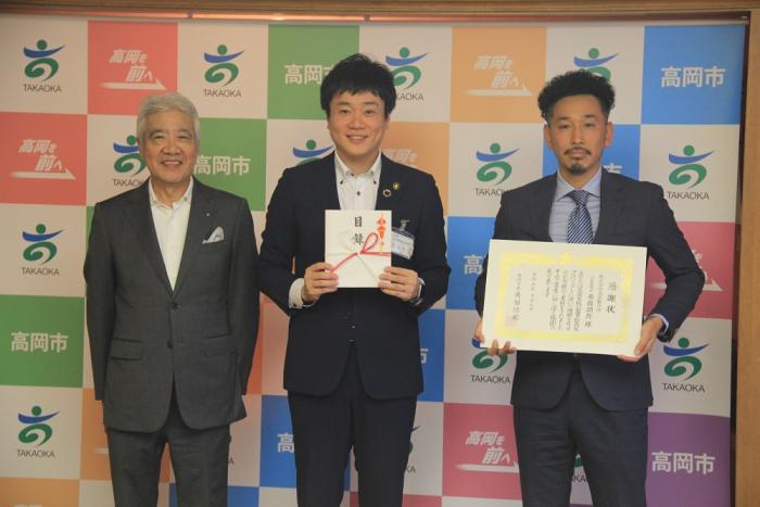 スーツを着た3人の男性が立って目録と感謝状を掲げている高岡製作所様の写真