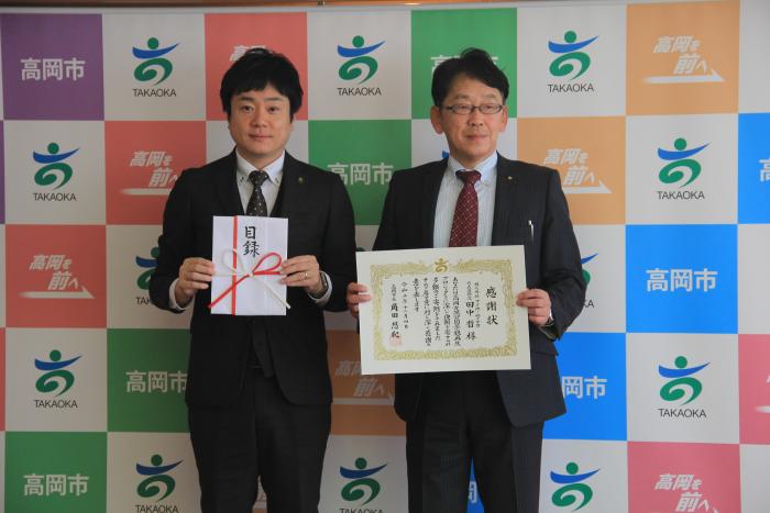 スーツを着た2人の男性が立って目録と感謝状を掲げている株式会社マイホームタナカ様の写真